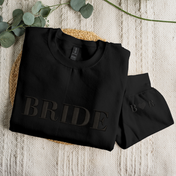 Black sweatshirt with 'BRIDE' text on a textured surface with eucalyptus leaves.