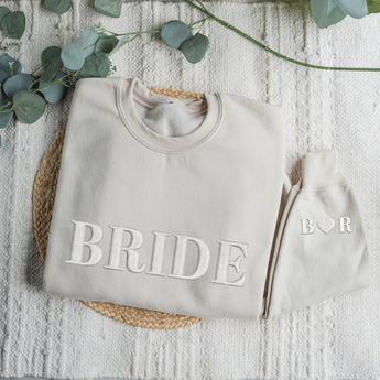 Gray sweatshirt with 'BRIDE' text and matching beanie on a textured surface with eucalyptus leaves.