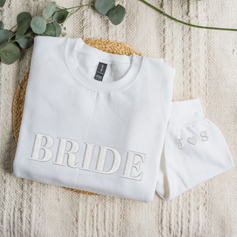 White sweatshirt with 'BRIDE' text on a textured surface with eucalyptus leaves.