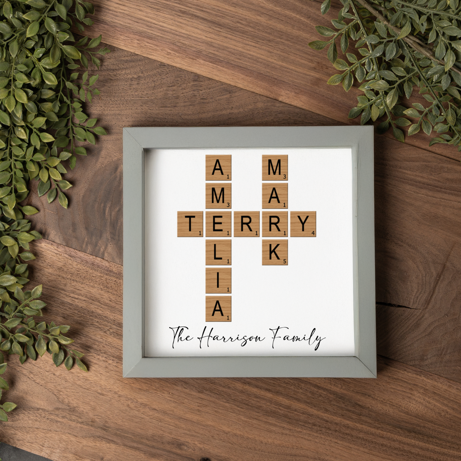 A personalized family name wall art piece featuring wooden scrabble-style tiles spelling “AMELIA,” “MARK,” and “TERRY” arranged in a crossword pattern on a white background. The phrase “The Harrison Family” is printed below in elegant black script. The artwork is displayed in a light gray frame on a warm walnut wood surface with greenery accents. A thoughtful custom family gift, perfect for home décor, anniversaries, and personalized gifts.