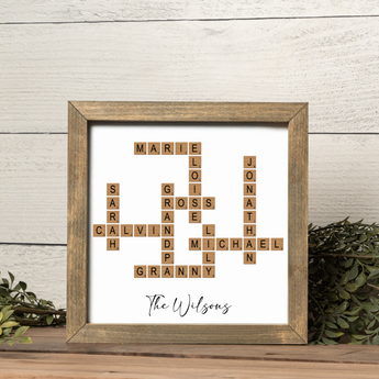 A custom crossword family name frame featuring scrabble-style wooden tiles arranged to spell multiple family names including “MARIE,” “CALVIN,” “JONATHAN,” and “GRANNY.” The text “The Wilsons” is elegantly written in black script beneath the design. Displayed in a rustic walnut-toned frame with greenery accents, this personalized family crossword art makes a unique home décor accent and a heartfelt gift for grandparents, parents, or family celebrations.