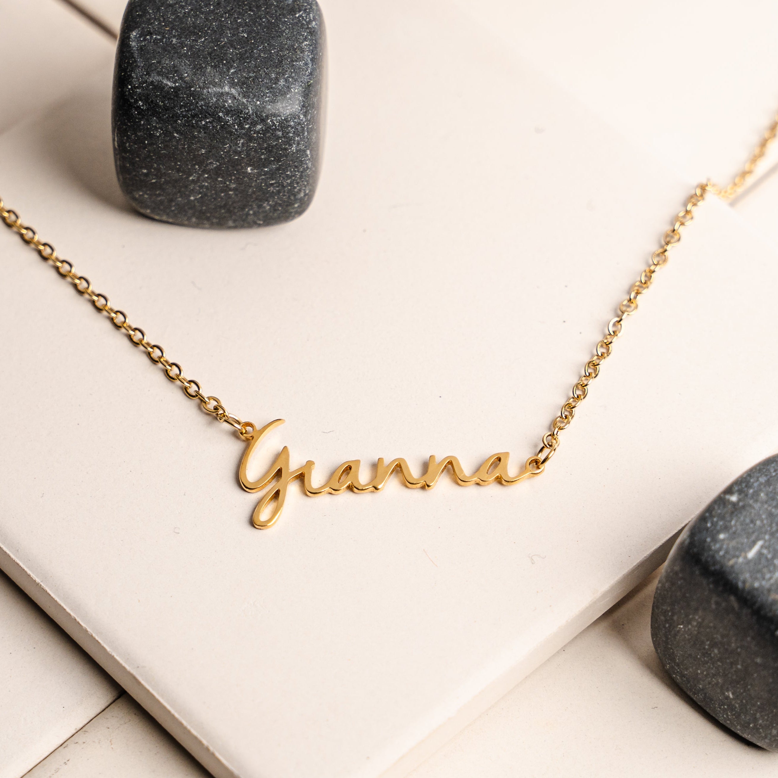 Gold necklace with 'Gianna' pendant on a white surface with gray stones.
