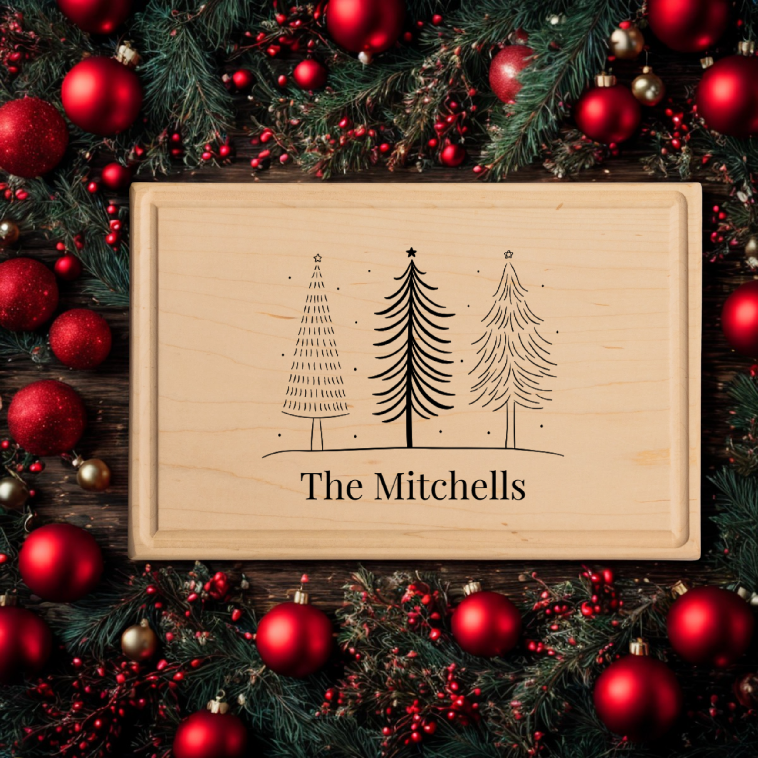 A personalized cutting board made from light maple wood engraved with three minimalist Christmas trees and the text “The Mitchells.” The board is surrounded by festive holiday greenery, red ornaments, and berries on a rustic wood background. This custom cutting board makes a perfect Christmas gift for families, newlyweds, or holiday hosts looking for personalized cutting board gifts.
