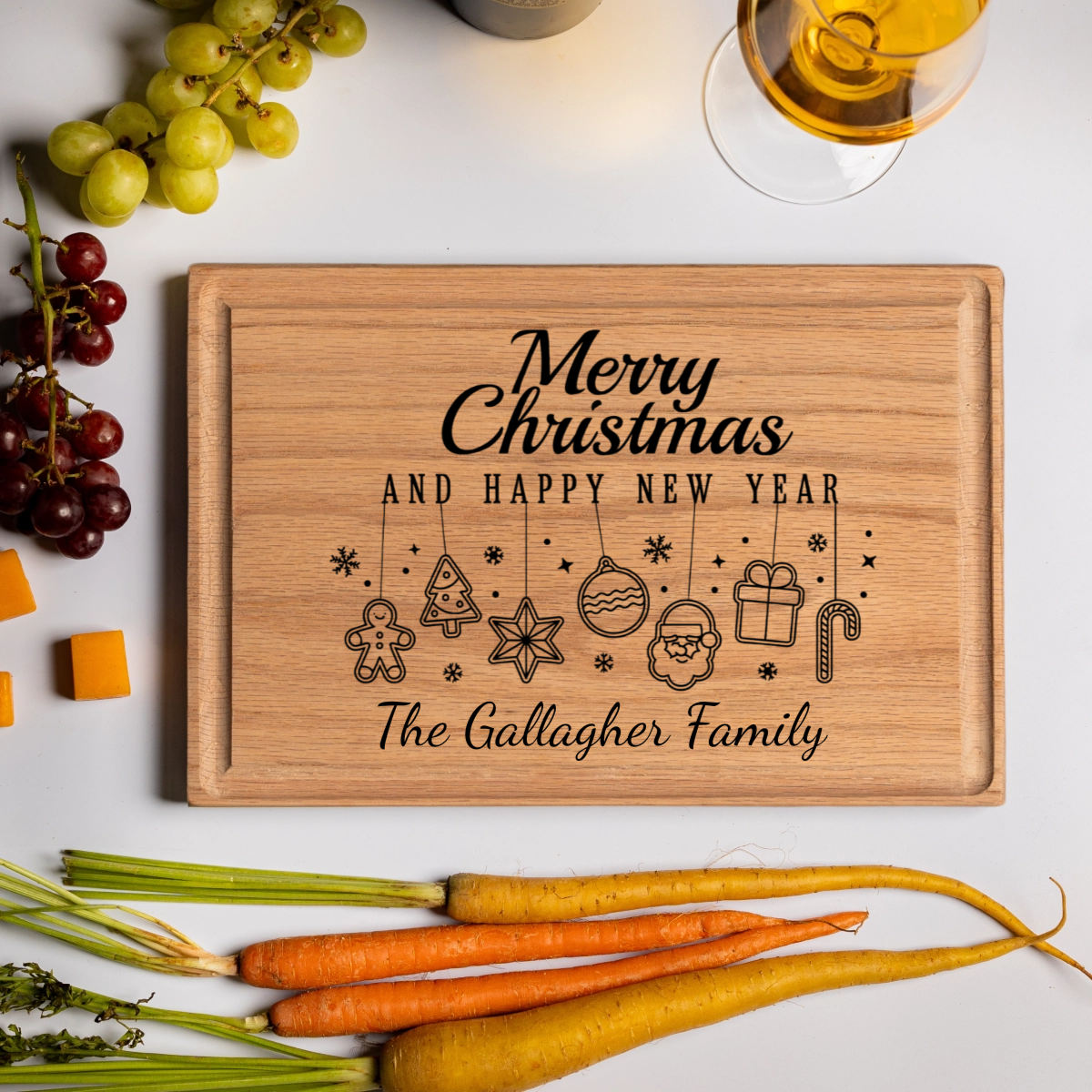 A personalized cutting board made from smooth oak wood engraved with “Merry Christmas and Happy New Year” above festive hanging icons including a gingerbread man, tree, star, ornament, Santa face, present, and candy cane. The board features “The Gallagher Family” in elegant script and is styled with grapes, cheddar cubes, a glass of golden wine, and colorful heirloom carrots. This custom cutting board is a beautiful Christmas gift idea and a functional holiday cutting board gift for families and hosts.